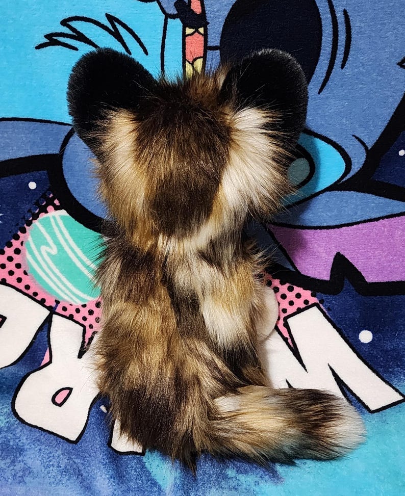 May include: A plush toy in the shape of a fox, featuring a mix of brown, tan, and black faux fur. The fox has large, black-tipped ears and a fluffy tail. The toy is set against a colorful, patterned background.