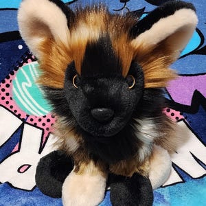 May include: A plush African wild dog toy with a black muzzle, ears, and paws. The toy has tan and brown fur around the face and body, with large, upright ears. The stuffed animal is sitting on a blue patterned surface.
