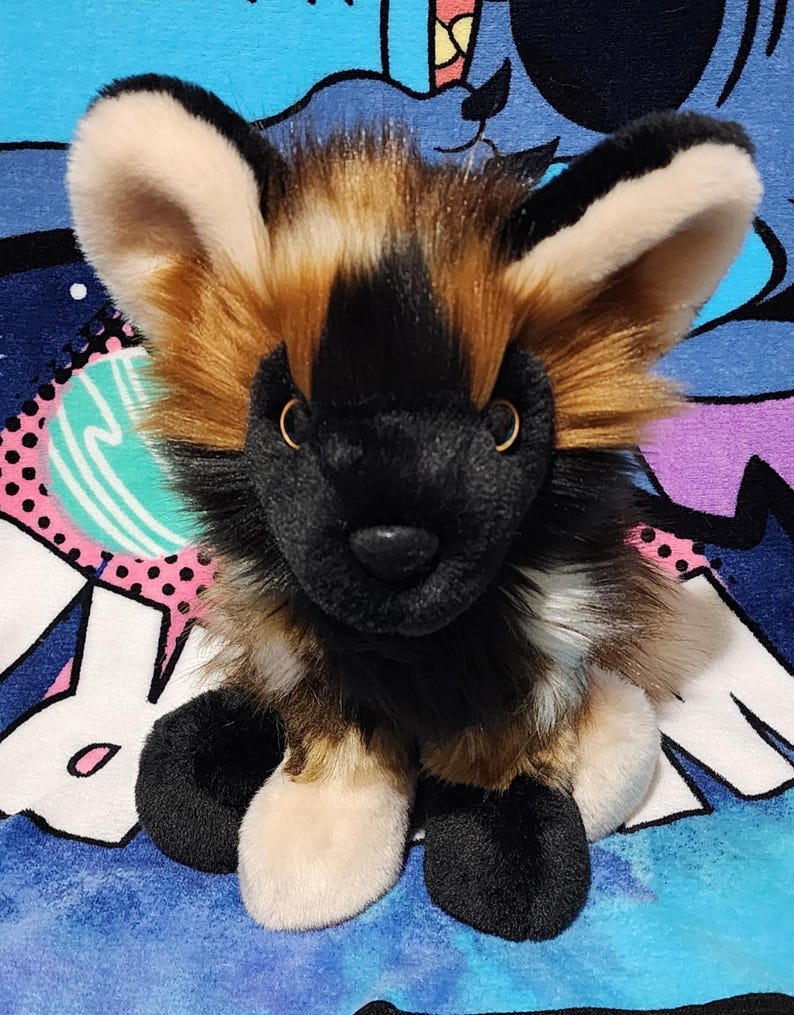 May include: A plush African wild dog toy with a black muzzle, ears, and paws. The toy has tan and brown fur around the face and body, with large, upright ears. The stuffed animal is sitting on a blue patterned surface.