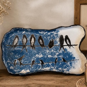 Peut inclure: Un plateau décoratif en céramique avec un fond bleu et blanc. Des oiseaux noirs sont perchés sur un fil, certains en vol. Le plateau a une forme irrégulière unique et un bord noir. Le plateau est sur une surface en bois.