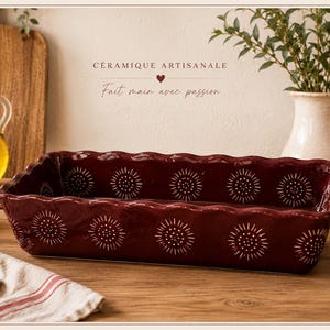 Könnte beinhalten: Eine rechteckige, bordeauxfarbene Keramik-Backform mit gewelltem Rand und weißen Sonnenmuster-Designs. Das Bild zeigt auch ein Schneidebrett, Olivenöl, Knoblauch und den Text "Céramique Artisanale, Fait main avec passion."