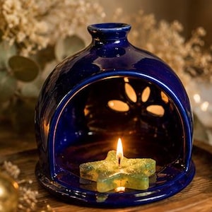 May include: A dark blue ceramic oil warmer with a star-shaped candle inside. The warmer has a dome shape with cut-out designs. A lit candle and a gold star are on a wooden tray, with a glass candle holder and decorative greenery in the background.