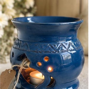 May include: A blue ceramic essential oil diffuser with a lit candle inside. The diffuser has decorative cutouts and a textured pattern around the middle. A small tag is attached with twine. The background is blurred with white flowers.