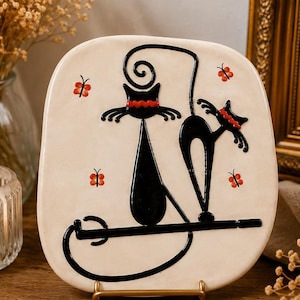 May include: A cream-coloured, square ceramic plate featuring a black cat design. Two stylised black cats are perched on a black line, adorned with red accents. Small red butterflies are scattered around the cats.