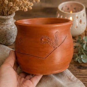 Peut inclure: Un pot en céramique de couleur terre cuite avec un large bord et un corps arrondi. Le pot présente des motifs floraux sculptés et une surface texturée. Le pot est probablement utilisé à des fins décoratives ou comme pot de fleurs.