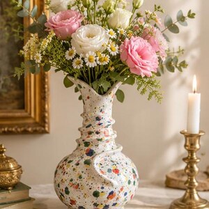 以下が含まれることがあります： 白の陶器の花瓶に、ピンクと白のバラ、デイジー、緑の葉のブーケが入っています。 金色の燭台に灯されたろうそくが近くにあり、暖かく居心地の良い雰囲気を作り出しています。