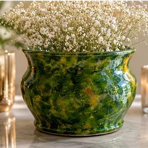 Puede incluir: Una maceta de cerámica vidriada verde y amarilla llena de flores blancas. La maceta redonda y decorativa tiene un acabado brillante y una amplia abertura. Una vela y un jarrón dorado están en el fondo.