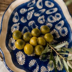 Può includere: Un piatto da portata in ceramica con un motivo floreale blu e bianco contiene olive verdi. Un bicchiere di liquido è sullo sfondo. Il piatto ha un bordo ondulato e un bordo color crema. Un rametto di foglie è a destra.