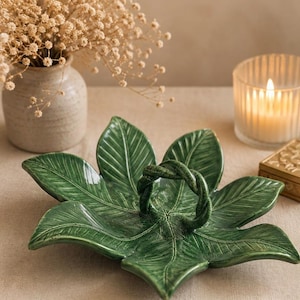Può includere: Un piatto da portata in ceramica verde a forma di foglia con un manico intrecciato. Il piatto presenta dettagliati motivi a venature di foglie ed è posto su una superficie neutra. Una candela accesa e un piccolo vaso con fiori secchi sono sullo sfondo.