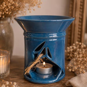 May include: A blue ceramic essential oil diffuser with leaf-shaped cutouts and a small tea light candle inside. The diffuser has a wide top and a cylindrical base, with a natural rope detail. The background includes dried flowers and a wooden surface.