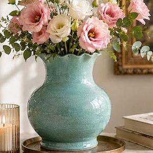 Könnte beinhalten: Eine türkisfarbene Keramikvase mit gerüschtem Rand enthält einen Strauß rosa und weißer Blumen mit grünem Laub. Die Vase steht auf einem goldfarbenen Tablett. Links ist eine brennende Kerze in einem Glashalter.