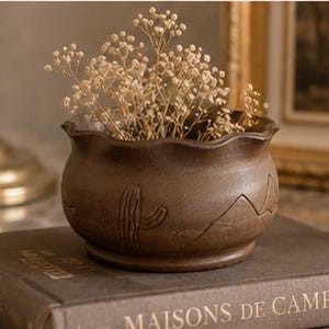 Op de afbeelding: Een kleine, bruine keramische pot met een golvende rand, met een uitgesneden cactusontwerp. De pot is gevuld met gedroogde, lichtgekleurde bloemen. Hij staat op een boek met de tekst "MAISONS DE CAMPAGNE".
