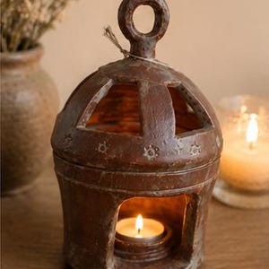 Pode incluir: Um castiçal rústico castanho com uma pega circular e recortes decorativos. Uma vela de chá acesa brilha no interior, projetando uma luz quente. O castiçal é feito de um material texturizado e é um detalhe decorativo para a casa.