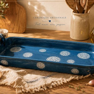 Peut inclure: Un plat à four rectangulaire en céramique bleu foncé avec des motifs floraux blancs. Il est doté de poignées en bois et repose sur un tissu en lin. L'image montre également de l'ail, une cuillère en bois et le texte "Céramique Artisanale, Fait main avec passion."