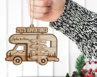 May include: A wooden camper ornament with the text "2025 ADVENTURES" and the names "Sophia & Henry". The ornament features directional signs pointing to various locations. The ornament is hanging from a gold chain.