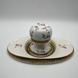 May include: A white ceramic inkwell set featuring a round ink pot and an oval base. Both pieces are decorated with small pink rose patterns and green leaves. The base and the lower section of the ink pot are finished with a gold-toned trim.