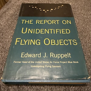 May include: A hardcover book titled "THE REPORT ON UNIDENTIFIED FLYING OBJECTS" by Edward J. Ruppelt. The book has a teal cover with yellow and white text. The author's role as former head of the United States Air Force Project Blue Book is also noted.