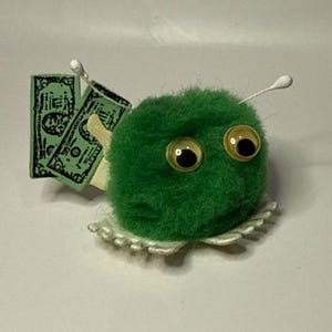 May include: A fuzzy green pom-pom creature with two large googly eyes and two white antennae. The creature features a white scalloped base and holds two rectangular green paper cutouts that resemble United States dollar bills behind its body.