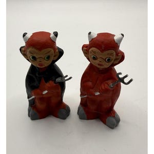 May include: Two small devil figurines with red skin, white horns, and grey hooves. One figure wears a black cape, while the other wears a red cape. Both figures have large eyes and hold a small, silver-colored metal pitchfork in their hands.