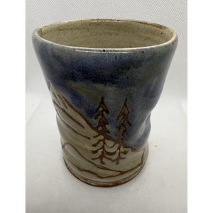 May include: Handmade ceramic tumbler with a landscape design. The tumbler features a blue and tan color scheme with a mountain and tree design. The tumbler is approximately 5 inches tall.