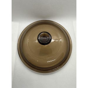 May include: A round, brown glass lid with a dark brown knob. The lid is slightly domed and has a transparent appearance. The knob is circular and appears to be made of the same material as the lid. This kitchenware item is designed to fit on a pot or pan.