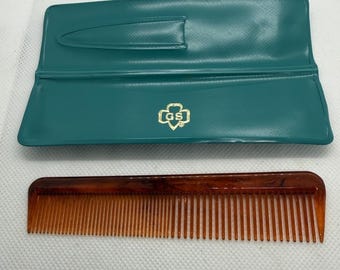 May include: A vintage comb and case set. The comb is a warm brown color with fine teeth. The case is teal green with a gold emblem. The case is open, revealing the comb.
