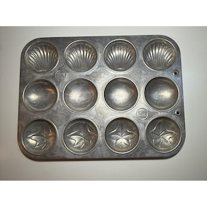 May include: A vintage, rectangular, aluminum baking pan with twelve molds. The molds are in the shape of seashells, circles, and leaves. The pan has a silver finish and is likely used for baking madeleines or other small pastries.