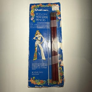 May include: Vintage Vogart Crafts iron-on transfer pencils for light and dark fabrics. The package is blue with floral accents and features an illustration of a woman in white pants. The pencils are a deep red color.