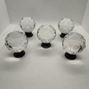 May include: Five clear crystal ball cabinet knobs with faceted surfaces. Each knob is mounted on a dark bronze-colored base. The knobs are arranged in a circular pattern, showcasing their reflective qualities.