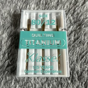 May include: A package of quilting needles in a clear plastic case. The needles are gold-colored with a size of 80/12. The package has a teal label with the word "TITANIUM" and the brand name "Klasse". The text "125 YEARS German Experience" is also visible.