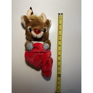 May include: A plush reindeer toy with a red nose, brown fur, and cream-colored antlers. The reindeer is holding a red blanket. The toy measures approximately 10 inches in height.