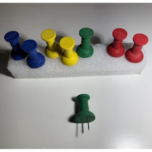 May include: A collection of colorful push pins in blue, yellow, green, and red. The push pins are arranged on a white foam base. One green push pin is isolated below the foam base.