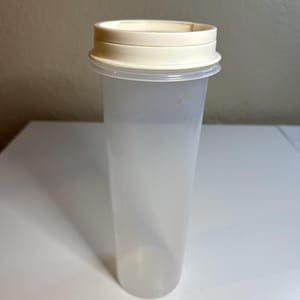 May include: A clear, cylindrical container with a cream-colored lid. The container is made of translucent plastic and has a simple, functional design. The lid appears to be made of a slightly opaque plastic. The container is empty.