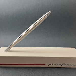 May include: A silver pen, inscribed with "pininfarina", rests on a cream-coloured box. The pen is angled, highlighting its streamlined design. The box features a red stripe and the "pininfarina" logo.
