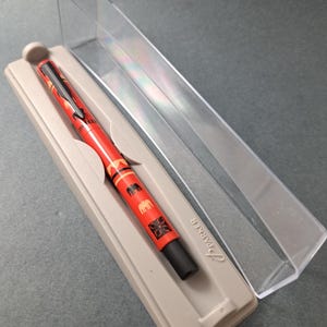 Puede incluir: Un bolígrafo rojo con detalles negros y un patrón de elefantes y formas geométricas, presentado en un estuche de plástico transparente. El bolígrafo está en un soporte beige. La marca "Parker" es visible.