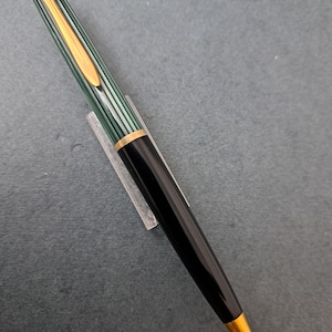 Puede incluir: Un bolígrafo a rayas negras y verdes con detalles dorados. El bolígrafo tiene una punta dorada y un cuerpo negro. El diseño incluye un clip dorado y una banda dorada cerca de la parte superior del bolígrafo.