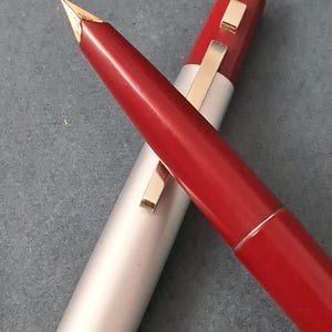 May include: A Pilot fountain pen with a red barrel and a silver cap. The pen features an integrated gold-coloured nib and a gold-coloured clip attached to the cap. The word PILOT is engraved in small letters near the bottom edge of the silver cap.