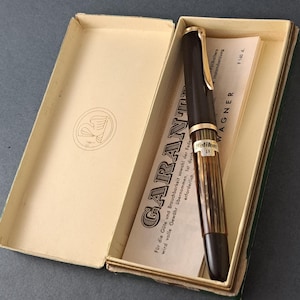 Puede incluir: Una pluma estilográfica vintage en una caja color crema. La pluma es de color marrón oscuro con detalles dorados y un cuerpo a rayas. La caja contiene una tarjeta de garantía con texto en alemán. La pluma está etiquetada como "Pelikan".