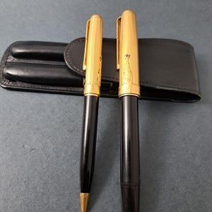 May include: A black and gold pen set comprising a ballpoint pen and a fountain pen, both featuring gold caps with vertical pinstripes and a small red star emblem. The pens are accompanied by a black leather case with two individual slots.