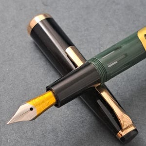 Puede incluir: Primer plano de dos plumas estilográficas. Una pluma es negra con detalles dorados y un plumín dorado. La otra pluma es de color verde oscuro con detalles dorados. El plumín de la pluma negra es visible, mostrando el intrincado diseño.