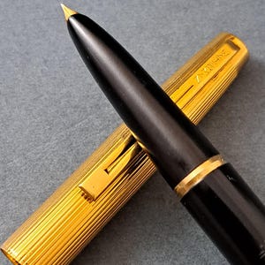 Peut inclure: Un stylo plume noir avec une plume et des détails dorés. Le capuchon doré présente un design rainuré. Le stylo est posé sur un fond gris.