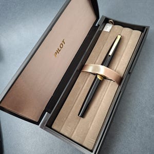 Könnte beinhalten: Ein schwarzer Pilot-Füllfederhalter mit goldfarbenen Akzenten liegt in einer rechteckigen Präsentationsbox. Die Box verfügt über einen Klappdeckel mit dem in Gold gedruckten Wort PILOT, ein beiges, samtgefüttertes Innere und ein Band.