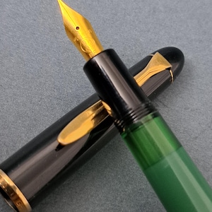 以下が含まれることがあります： 金色のペン先と黒と緑のボディの万年筆のクローズアップ。ペンのキャップは開いており、ペン先が見えています。このペンは金色のアクセントと洗練されたデザインで、筆記やコレクションに適しています。