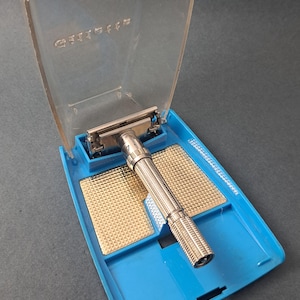 Puede incluir: Afeitadora de seguridad Gillette vintage en un estuche de plástico azul con tapa transparente. La afeitadora tiene un mango y una cuchilla plateados. El estuche tiene inserciones doradas. La palabra "Gillette" está grabada en la tapa.