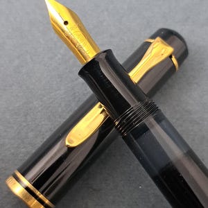 Könnte beinhalten: Ein schwarzer Füllfederhalter mit goldenen Akzenten, einschließlich Feder und Clip. Die Kappe ist abgenommen und zeigt die goldene Feder. Der Stift liegt auf einer grauen Oberfläche.