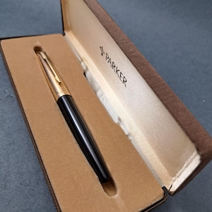 May include: A black and gold Parker pen in its original brown case. The pen is displayed open, revealing the pen nestled in a tan interior. The Parker logo is visible on the inside of the case.