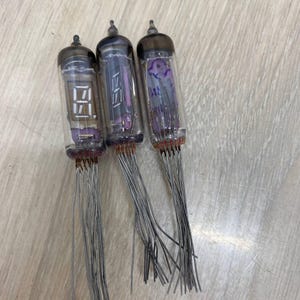 Może przedstawiać: Trzy zabytkowe lampy Nixie z szklanymi obudowami, każda z ciemną górą i licznymi przewodami. Lampy wyświetlają cyfry cyfrowe i elementy wewnętrzne w odcieniach fioletu i pomarańczy.