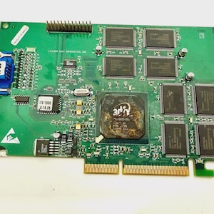 Puede incluir: Una placa de circuito verde con varios componentes electrónicos, incluyendo chips, conectores y un puerto VGA azul. La placa tiene texto y marcas, y un chip central grande con el logotipo "3dfx".