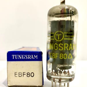 Peut inclure: Un tube à vide Tungsram EBF80, avec un boîtier en verre et des broches métalliques, à côté de sa boîte d'origine bleue et blanche. La boîte affiche le nom de la marque et le modèle du tube. Le tube est en verre transparent.