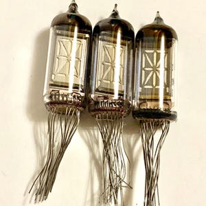 Może przedstawiać: Trzy zabytkowe lampy Nixie ze szklanymi obudowami i metalowymi pinami. Każda lampa ma ciemną, zaokrągloną górę i wyświetla segmentowy wyświetlacz cyfrowy. Rurki są ułożone pionowo na białym tle, prezentując swój unikalny design.
